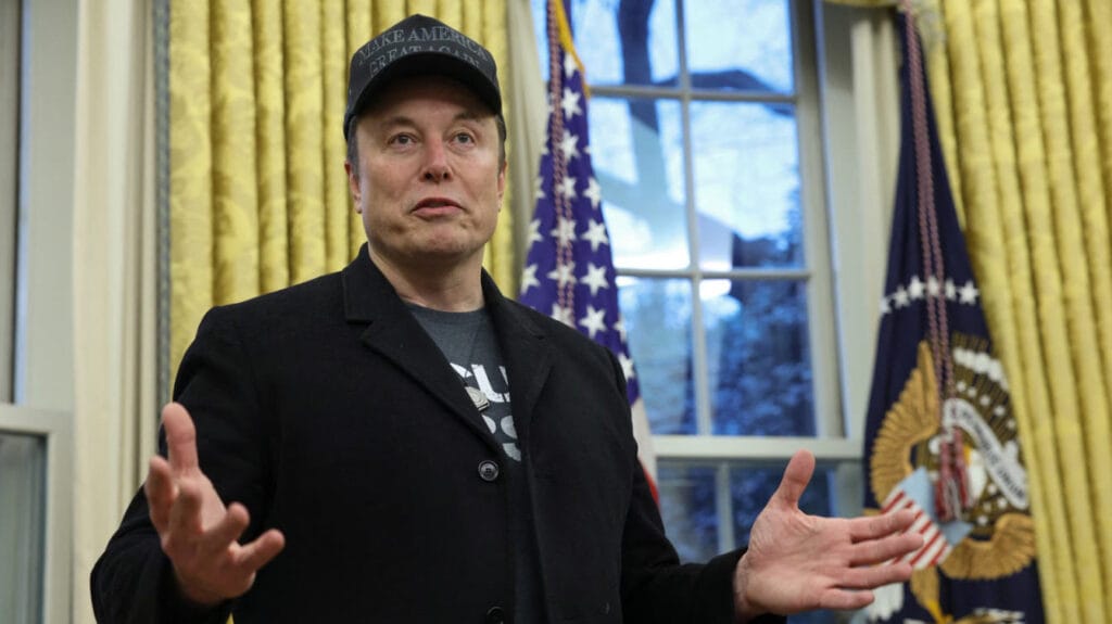 Elon Musk standing in the Oval Office wearing a "Make America Great Again" hat, with an American flag behind him.