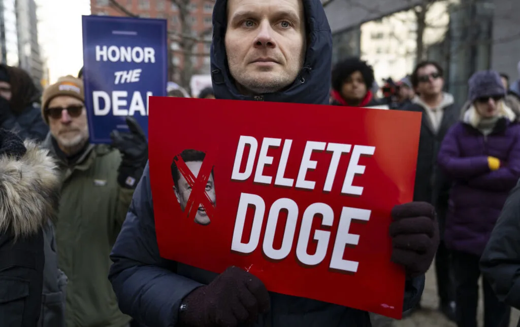 A protester in New York City holds a red sign that reads "DELETE DOGE" with a crossed-out image of Elon Musk, reflecting public opposition to the DOGE government restructuring efforts.