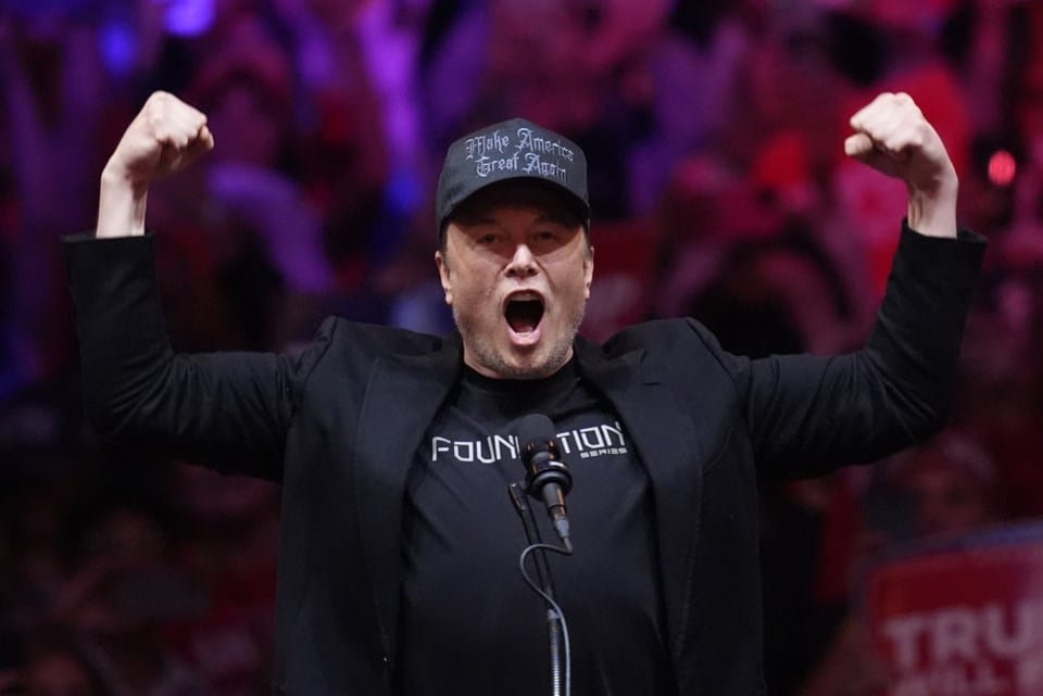 Elon Musk, dressed in black with a black "Make America Great Again" hat, raises his fists while speaking at a Trump rally, embracing the label of "Dark, Gothic MAGA and suggesting a connection to the Dark Enlightenment.