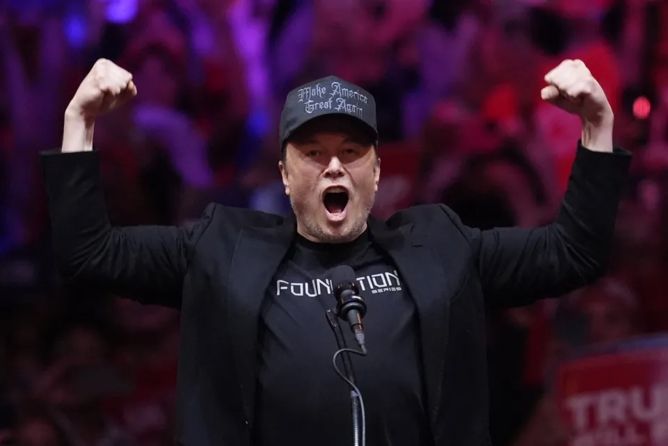 Elon Musk, dressed in black with a black "Make America Great Again" hat, raises his fists while speaking at a Trump rally, embracing the label of "Dark, Gothic MAGA and suggesting a connection to the Dark Enlightenment.