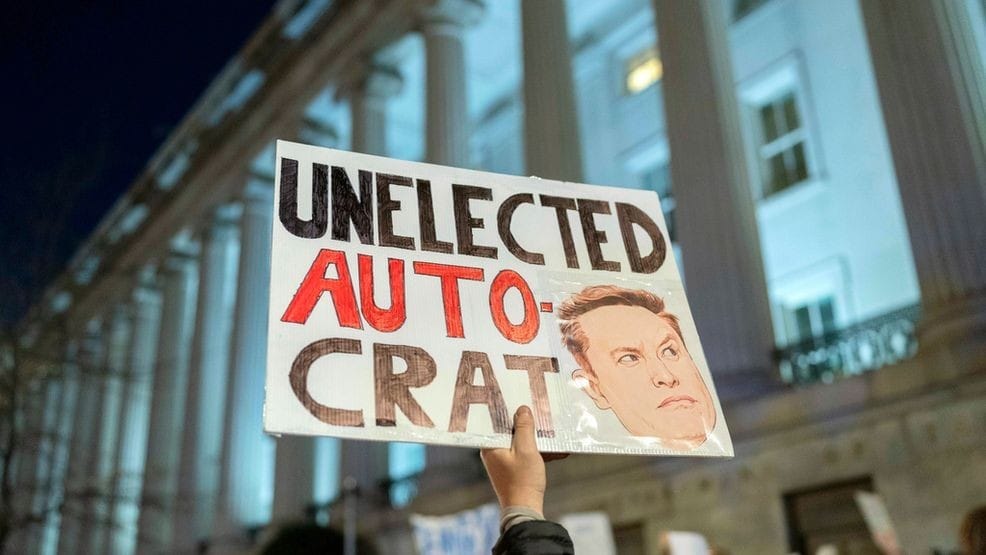 A protest sign reading "Unelected Autocrat" with an illustration of Elon Musk is held up outside the U.S. Treasury Department during a demonstration.